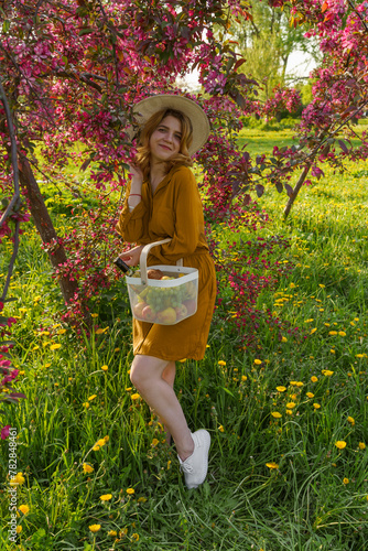 blonde girl stands in park against backdrop of blossoming tree, wearing hat, brown dress. She holds iron basket filled with fruits. girl is joyful, smiling, and basking in sunshine, summer vibes © Маргарита Трушина