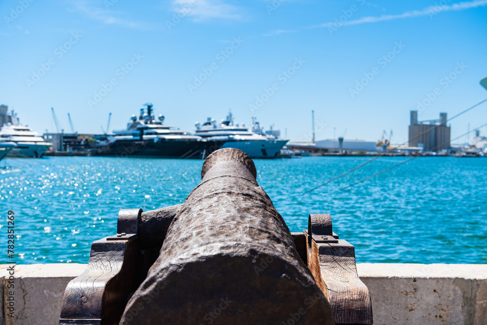 Eine alte Kanone im Hafen, im Hintergrund liegen Luxusyachten vor Anker, Tarragona, Spanien - obrazy, fototapety, plakaty 
