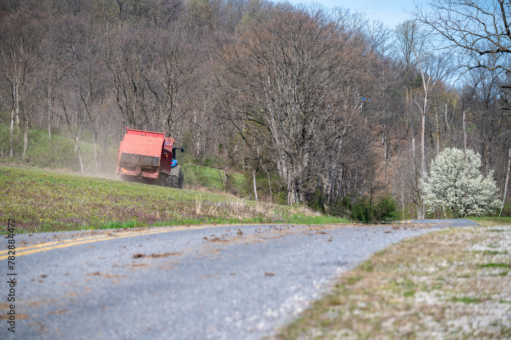 Farm tractor fertilizes agricultural field with red manure spreader next to a rural country road with no people and foreground defocused copy space