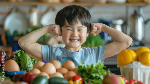 A young boy is standing in front of a table full of fruits and vegetables. He is smiling and flexing his arms muscle, eating healthy food concept