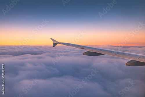 Airplane flying over color sky clouds during scenic sunset or sunrise cloudscape, view from plane window of wing turbines and horizon