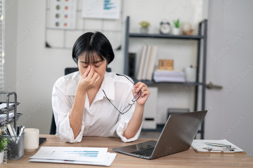Exhausted female office worker feeling stressed, holding glasses, with laptop and documents on desk. Exhausted female office worker feeling stressed, holding glasses, with laptop and documents on desk.