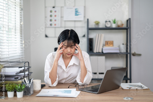 Exhausted female office worker feeling stressed, holding glasses, with laptop and documents on desk. © Wasana