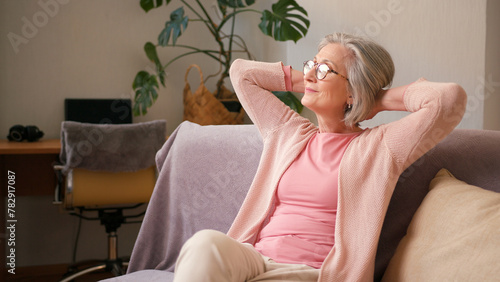 Mature old 60s woman, older middle aged female resting on couch taking deep breath of fresh air holding hands behind head at home