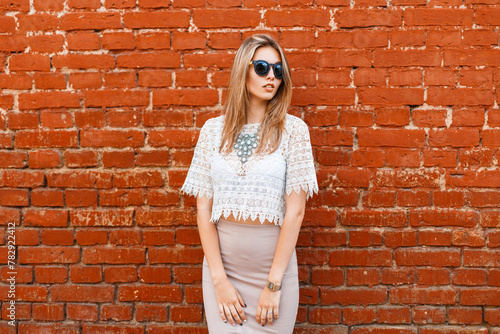 Beautiful young hipster woman with sunglasses in a white lace top with a skirt and jewelry stands near a red brick wall