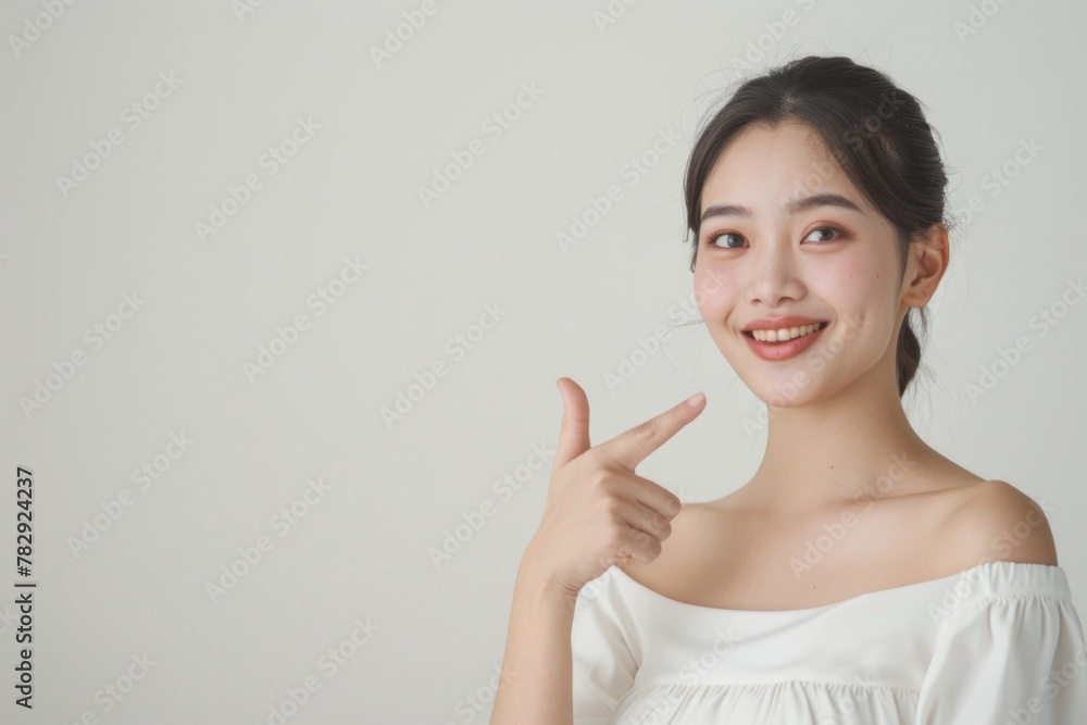Portrait of a beautiful smiling Asian woman in a white dress, pointing her finger up to the side on an empty light background. Portrait of a beautiful smiling Asian woman in a white dress, pointing her finger up to the side on an empty light background.