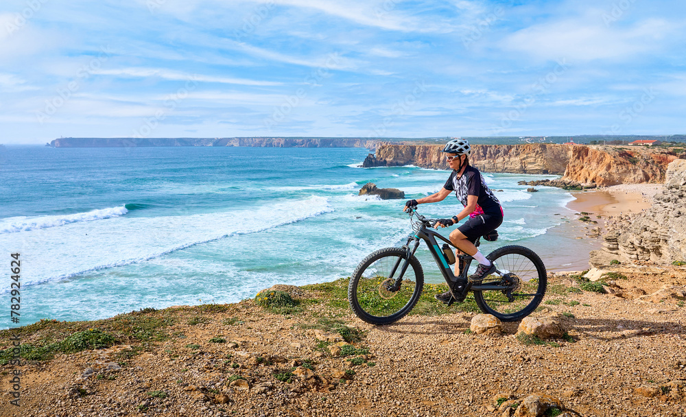 Fototapeta premium happy active senior woman cycling at the the rock cliffs and lighthouse of Cabo Sao Vicente, the south-western spit of Europe at the atlantic coast of Algarve, Portugal, 