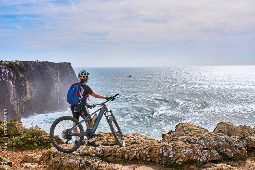 Obraz premium happy active senior woman cycling at the the rock cliffs and lighthouse of Cabo Sao Vicente, the south-western spit of Europe at the atlantic coast of Algarve, Portugal, 