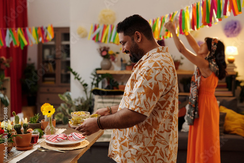 Side view of happy Hispanic man in Mexican shirt bringing homemade snacks for festive table against his girlfriend decorating living room © pressmaster