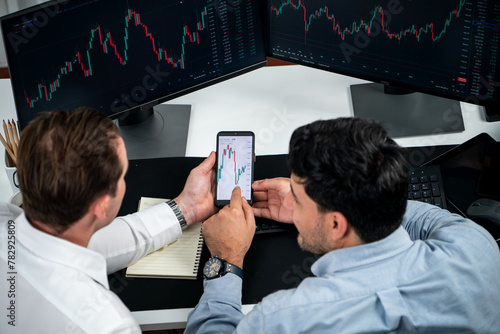 Investor stock officer focusing on dynamic exchange rate database on smartphone, comparing with market dynamic graph with monitor screen. Concept of analysis trading technology investment. Sellable. photo