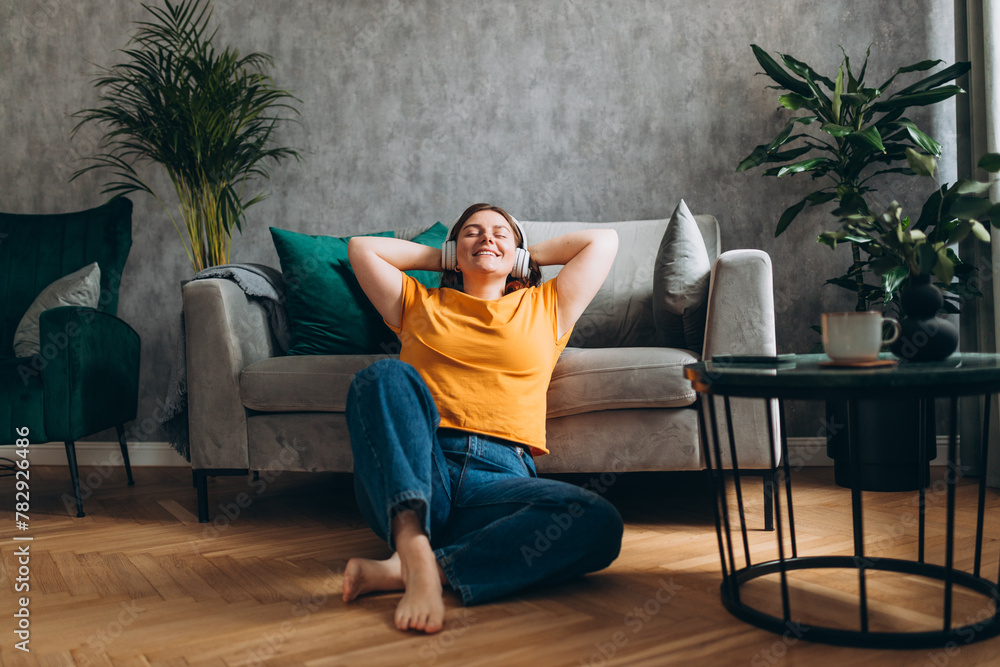 Smiling girl relaxing at home, she is playing music using a smartphone and wearing white headphones. Young woman hearing enjoying good audio sound lounge near couch at home, full body