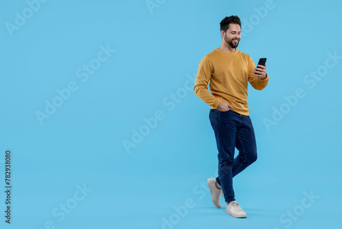 Happy young man using smartphone on light blue background, space for text photo