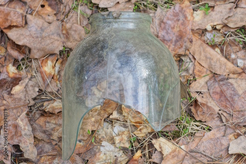one large gray broken glass jar with a hole lies on the ground in brown dry leaves on the street