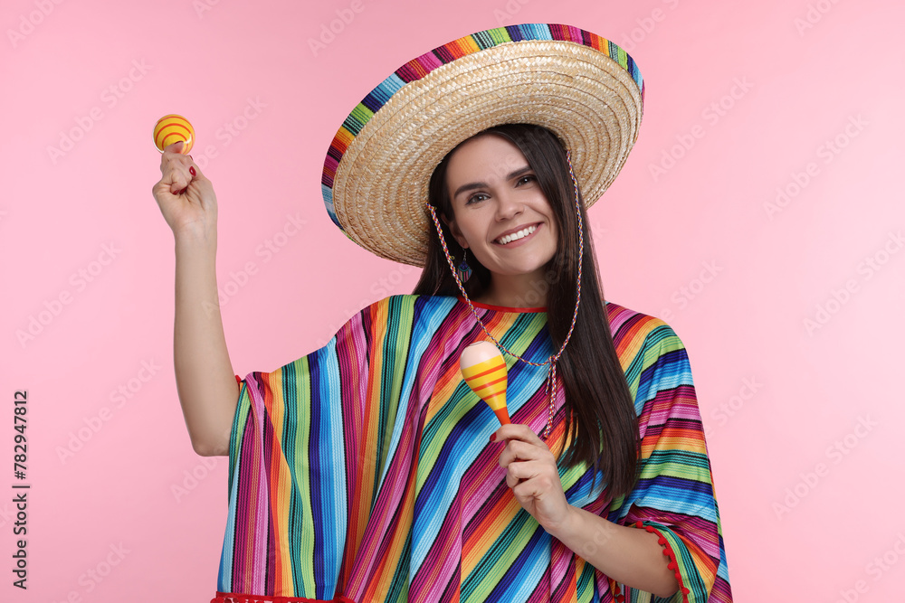 Young woman in Mexican sombrero hat and poncho dancing with maracas on pink background Young woman in Mexican sombrero hat and poncho dancing with maracas on pink background