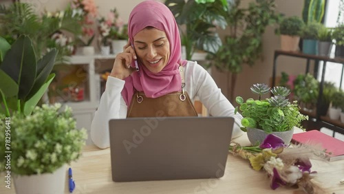 A smiling woman in a hijab talks on the phone while using a laptop in a plant-filled home office. photo