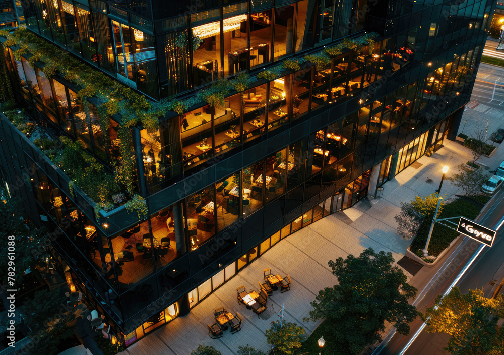 Aerial view of an urban hotel restaurant with large glass windows and outdoor seating, surrounded by contemporary architecture in the city center at night