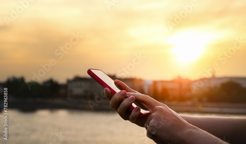 Female person touching screen of her phone with her finger in front of background of city during sunset. 