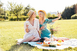Two young beautiful smiling hipster female in summer sundress and hats. Carefree women making picnic outside. Positive models sitting on plaid on grass, drinking champagne, eating fruits, take selfie