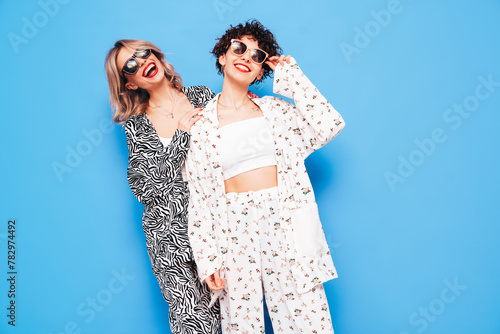 Two young beautiful smiling brunette hipster female in trendy summer costume clothes. Sexy women posing near blue wall in studio. Positive models having fun. Cheerful and happy. In sunglasses
