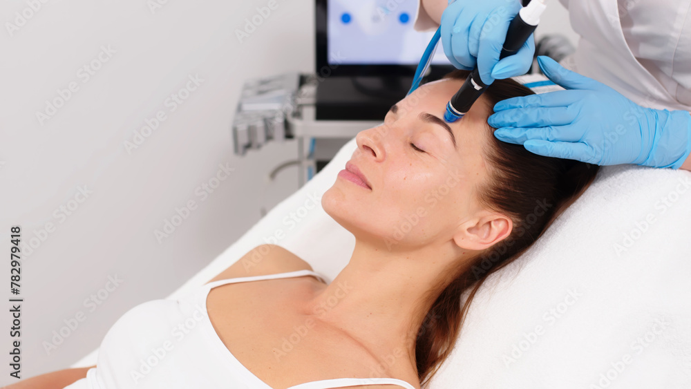 Fototapeta premium Close-up of a cosmetologist's hands polishing and cleansing the skin of a middle-aged woman's face using a vacuum machine. Concept of skin care, rejuvenation and cosmetology.