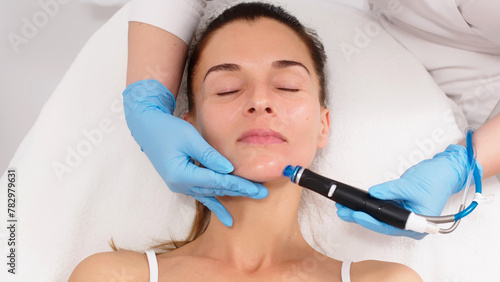A middle-aged woman receives facial treatment in a professional beauty salon. A close-up cosmetologist manipulates a hydropeeling device to clean and rejuvenate facial skin.
