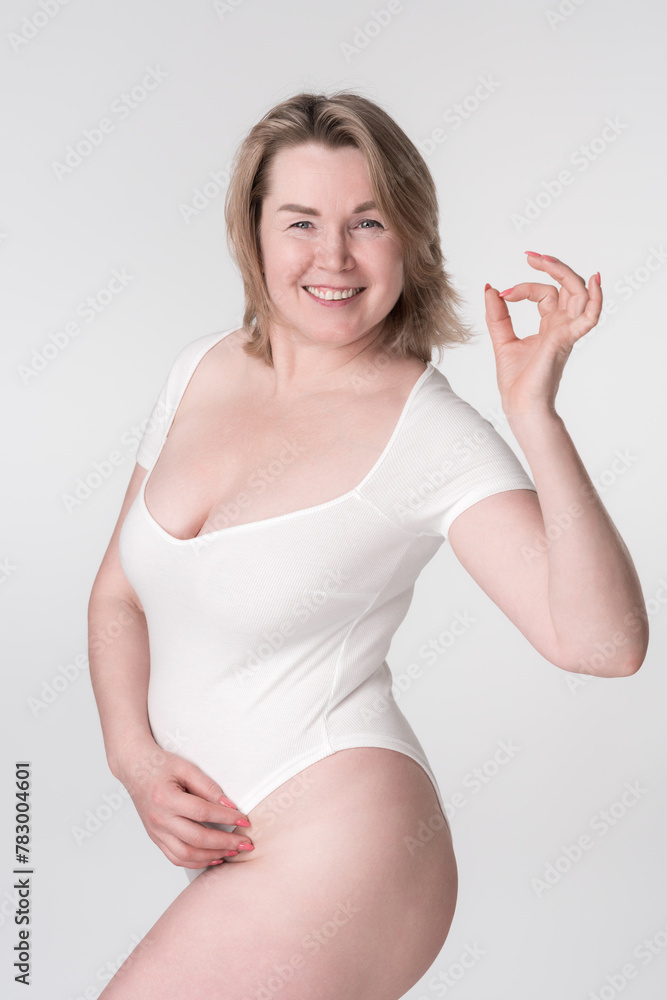 Happiness plus size woman in white bodysuit shows OK sign. Extended sizes mature adult model smiling and looking at camera posing on white background. Generation X curvy fashion model. Body positive. Happiness plus size woman in white bodysuit shows OK sign. Extended sizes mature adult model smiling and looking at camera posing on white background. Generation X curvy fashion model. Body positive.