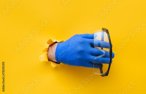 The male hand in a blue fabric work glove holds safety glasses. Torn hole in yellow paper. Good job, eye protection and safety concept. Copy space. photo