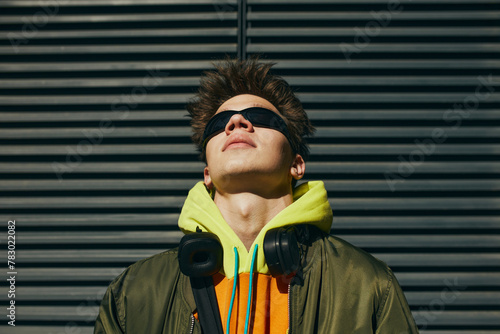 Portrait of man in trendy streetwear, neon hoodie, sunglasses, headphones posing against metal slatted background. Concept of street fashion and urban style, modern lifestyle, gen Z, self-expression.