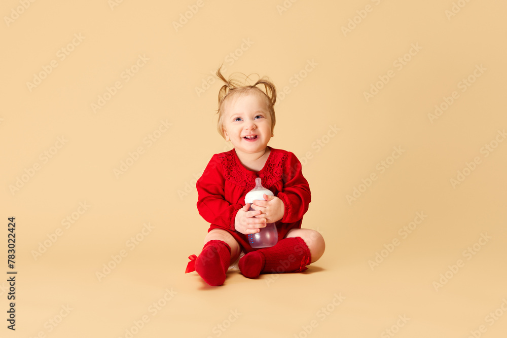 Little baby girl, child with ponytails in red dress and socks holding bottle, smiling, sitting on beige background. Concept of childhood, care, health, well-being, parenthood Little baby girl, child with ponytails in red dress and socks holding bottle, smiling, sitting on beige background. Concept of childhood, care, health, well-being, parenthood
