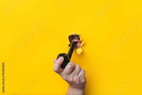 The hand of a young man holds a smoking pipe on a background of yellow paper. Mouth and lips in a ragged hole. Concept of bad habit and ancient way of smoking tobacco.