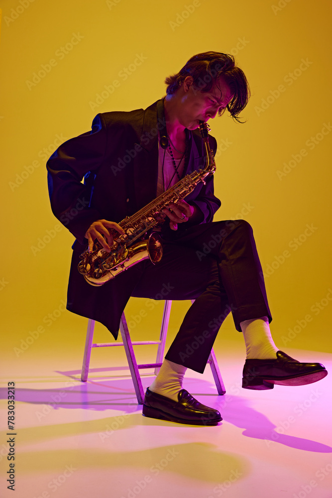 Fototapeta premium Young man, talented, charismatic solo player in black suit playing sax in neon light against vibrant yellow studio background. Concept of music and art, hobby, concerts and festivals, modern culture.