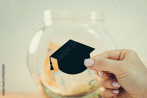 Black students hat made of paper, close up in female hand, toned photo, glass jar on the background. Concept of costs for education, saving money, investing in knowledge and profession © Vitalii