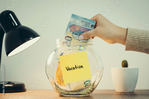 Twenty euro banknote in female hand, transparent glass money box with euros inside and word "Vacation" on it, toned. Long term investations, savings for free time, pleasure and relax
