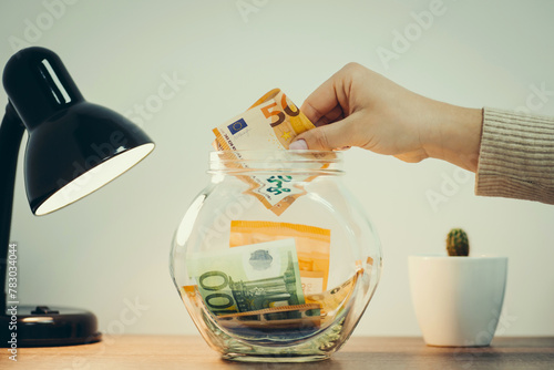 Money in the glass jar standing on the table, female hand putting 50 euro banknote inside, close up, toned picture. Saving funds for future, investments and finances concept