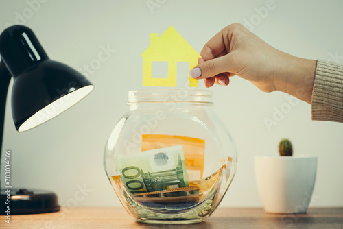 Close up shot of glass jar standing on the table with euro banknotes inside, female hand holding picture of a house above it. Toned photo with concept of saving funds for buying a house