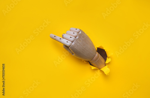 A wooden hand protrudes from a torn hole in yellow paper and points its index finger upwards to the left. Concept of direction, adjustment and assistance. Mannequin, robot