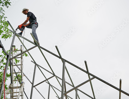 Man engineer standing on metal rail and using electric drill while installing support structure for photovoltaic solar panels. Isolated on white background.