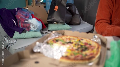 Handsome bearded man sitting in hoarder clutter and household junk in messy living room, working on laptop computer.