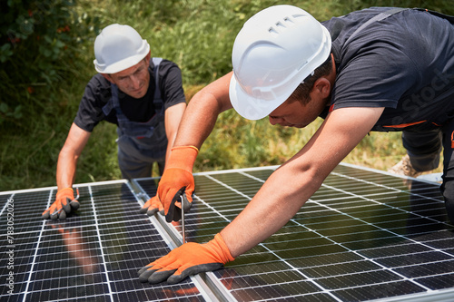 Male workers mounting photovoltaic solar panel system outdoors. Men engineers placing solar module on metal rails, wearing construction helmets and work gloves. Renewable and ecological energy. © anatoliy_gleb