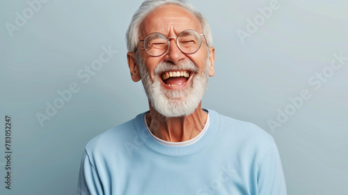 A man with a big smile on his face is wearing glasses and a blue shirt. He is laughing and he is happy. laughing old man, beard, glasses, happy, open laugh, enthusiastic, light blue sweater