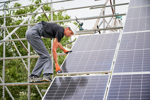 Worker building solar panels on metal beams. Man in helmet and uniform. Photo-voltaic collection of modules. Array as a system of photo-voltaic panels. Concept of renewable and ecological energy.
