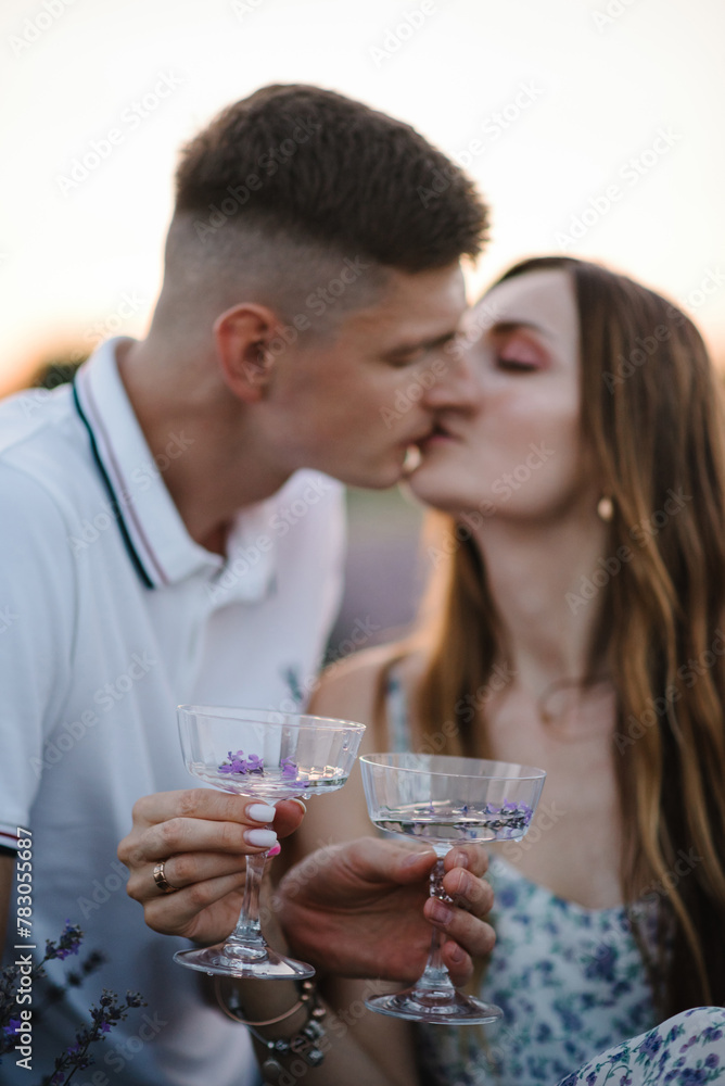 Male kisses female. Man hugs woman holds glasses of wine with lavender sprigs and drinks. Couple sits in lavender field on romantic evening at sunset. Straw basket and croissants on summer picnic. Male kisses female. Man hugs woman holds glasses of wine with lavender sprigs and drinks. Couple sits in lavender field on romantic evening at sunset. Straw basket and croissants on summer picnic.