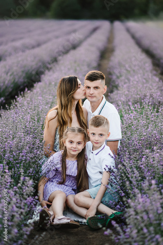 Mom kisses dad, son, daughter embrace, walk together in lavender field at sunset. Family vacation on summer day. Young mother, father, and two children hugging and sitting in purple lavender flowers.