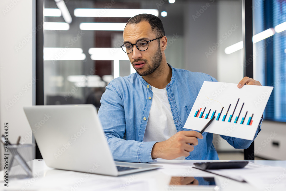 A young Latin American man works in the office, sits at a table in glasses, shows documents with graphs on a laptop camera, conducts an online meeting A young Latin American man works in the office, sits at a table in glasses, shows documents with graphs on a laptop camera, conducts an online meeting