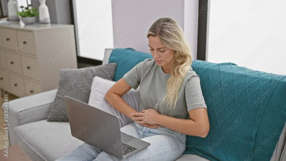 Young blonde woman experiencing stomach pain while working on her laptop in the living room of her home. Young blonde woman experiencing stomach pain while working on her laptop in the living room of her home.