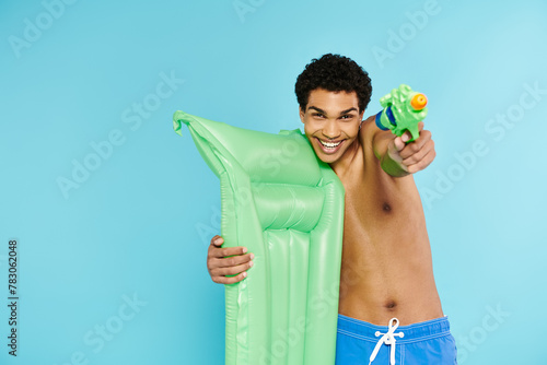 cheerful young african american man posing with air mattress and water gun on blue background