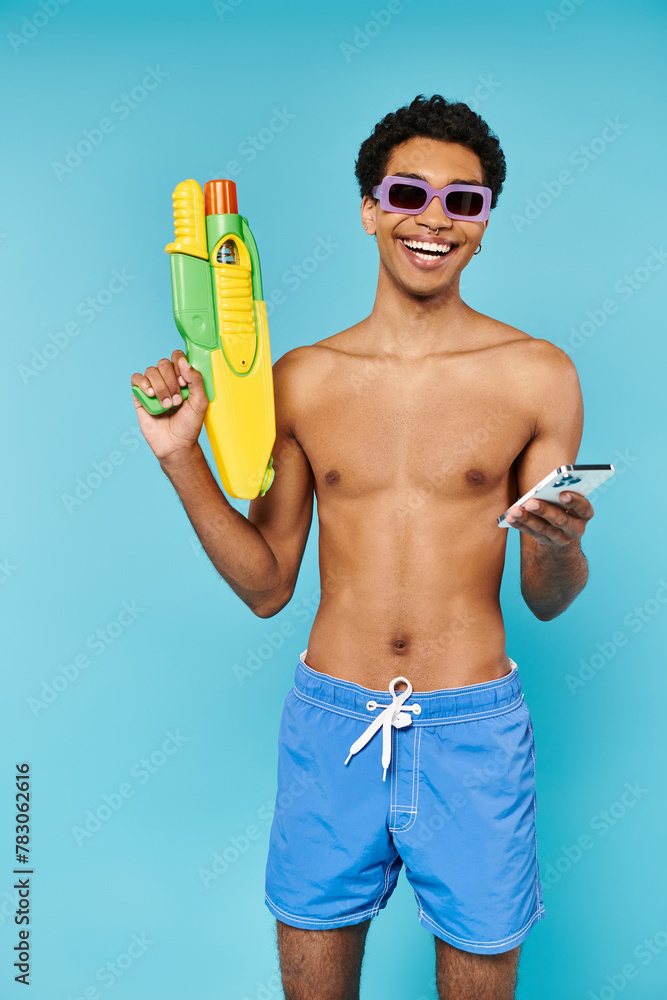 joyous african american man with stylish sunglasses holding phone and water gun on blue backdrop joyous african american man with stylish sunglasses holding phone and water gun on blue backdrop