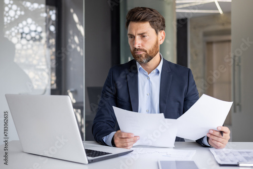 Bilde på lerret A serious young man businessman works in the office at a desk, looks at the scre