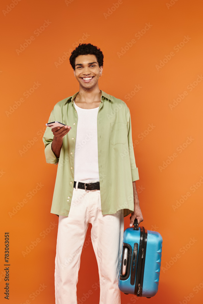 young cheerful african american man in cozy attire holding phone and suitcase and looking at camera young cheerful african american man in cozy attire holding phone and suitcase and looking at camera