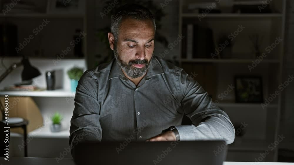 Focused mature hispanic man with beard and grey hair working late in a dark office Focused mature hispanic man with beard and grey hair working late in a dark office